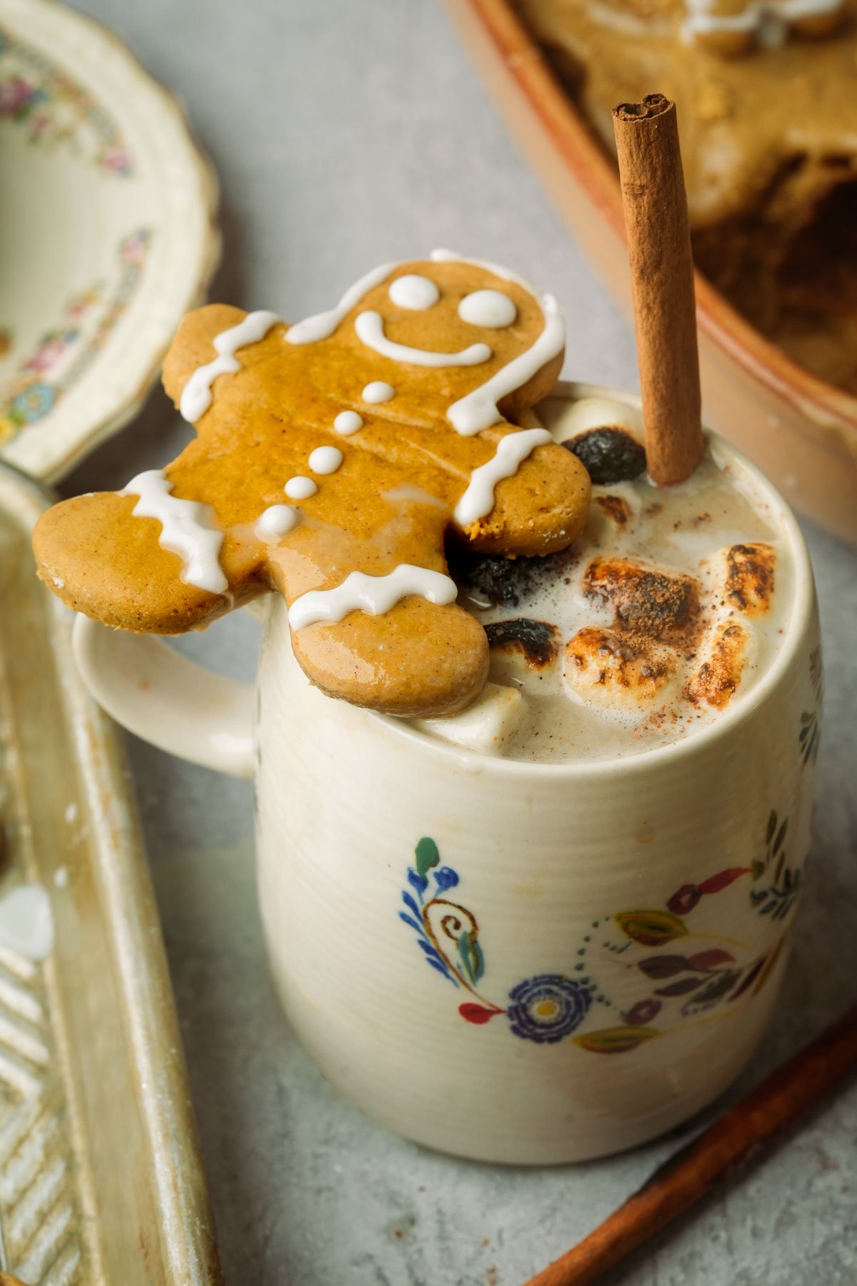 A mug of gingerbread latte topped with torched marshmallows, nutmeg, and a cinnamon stick, with a smiling gingerbread cookie perched on the rim. The mug features a colorful floral design.