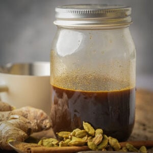A glass jar filled with dark brown gingerbread simple syrup sits on a surface next to fresh ginger, green cardamom pods, and a cinnamon stick, with a blurred white cup in the background.