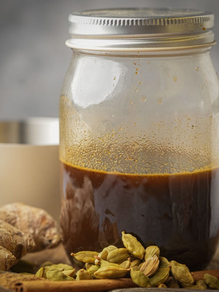A glass jar filled with dark brown gingerbread simple syrup sits on a surface next to fresh ginger, green cardamom pods, and a cinnamon stick, with a blurred white cup in the background.
