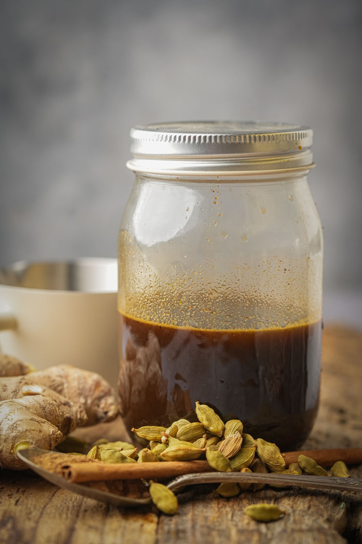 A glass jar filled halfway with dark brown gingerbread simple syrup and sealed with a metal lid sits on a wooden surface, surrounded by green cardamom pods, a piece of ginger root, and a metal spoon. A blurred cup is in the background.