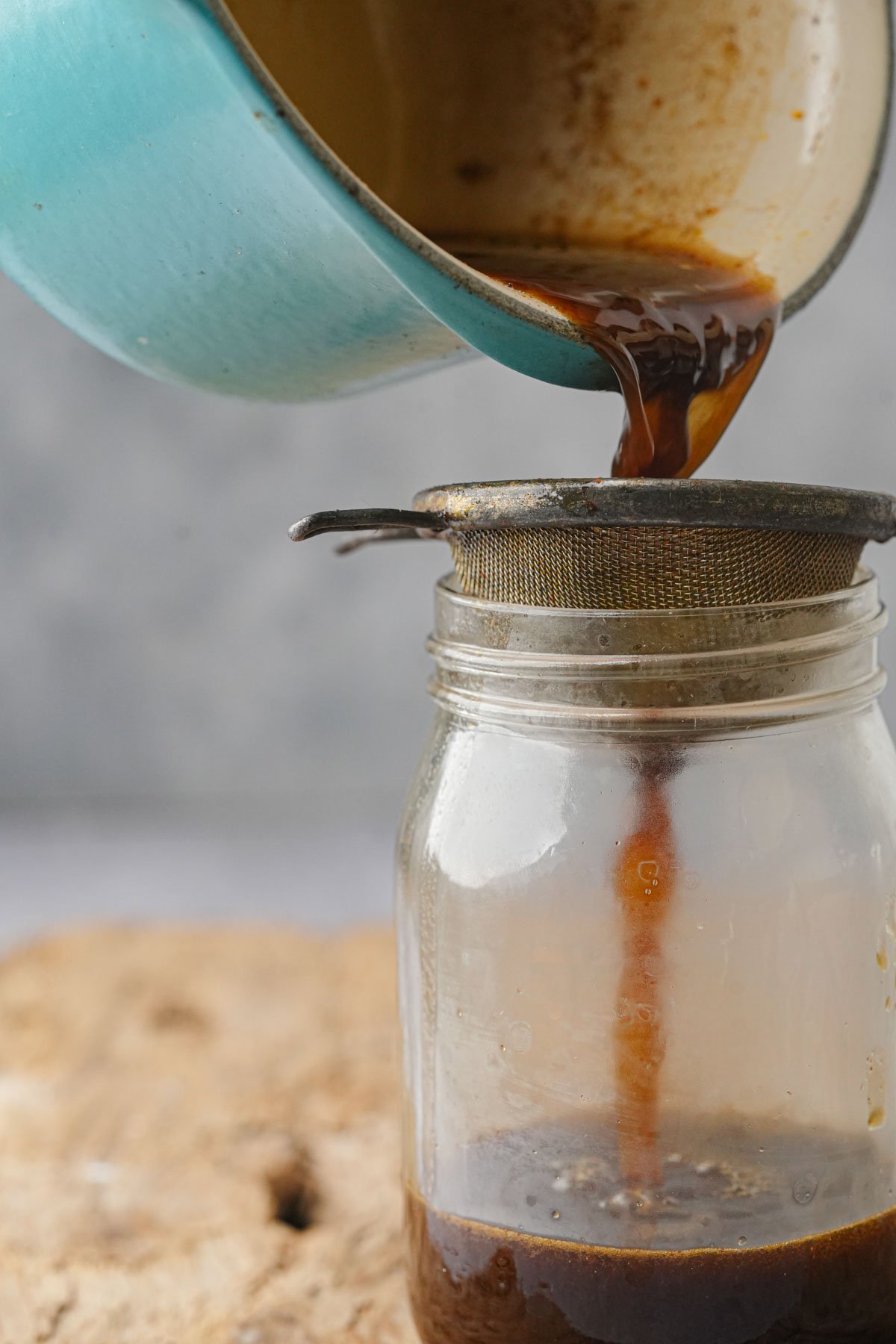 A blue pot pours cooled gingerbread simple syrup through a metal strainer into a glass jar, which is placed on a wooden surface.