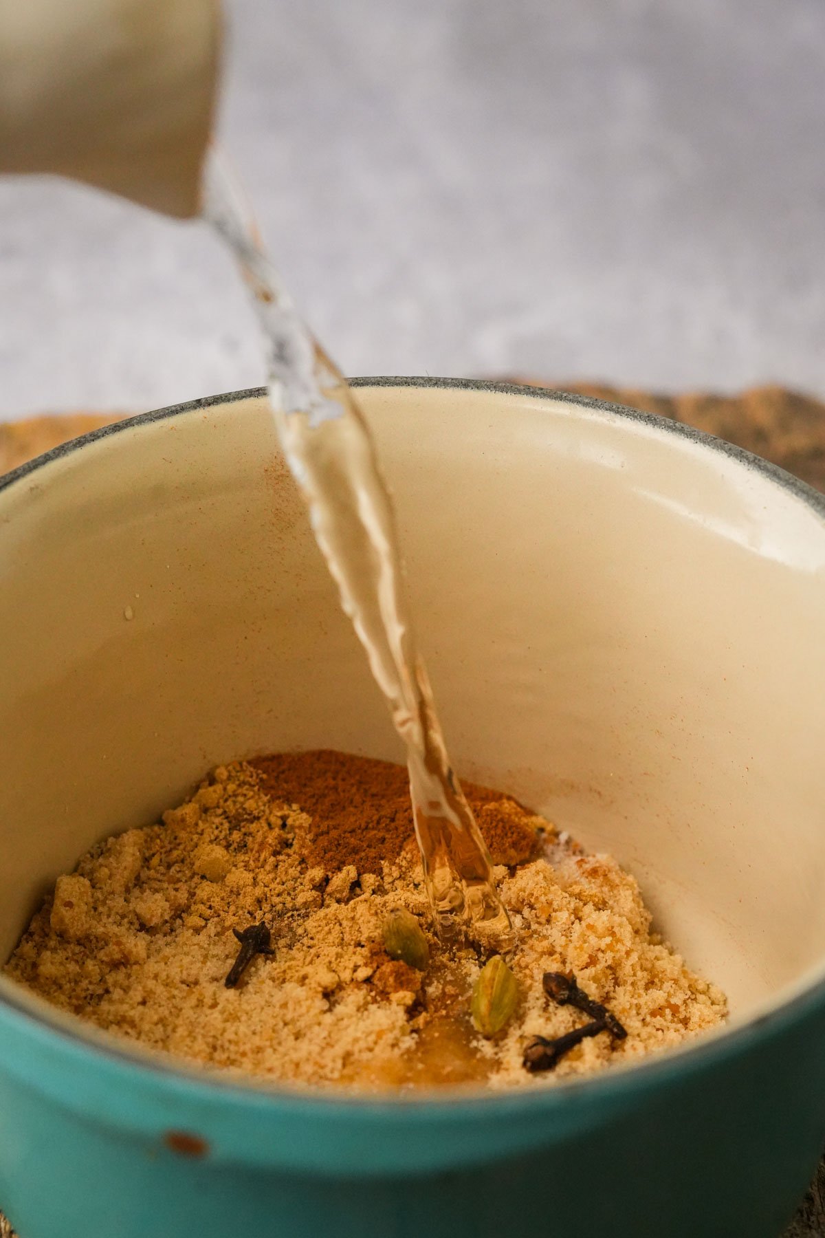 A stream of water is being poured into a blue ceramic bowl containing a mix of spices and powders, including cloves and cardamom, on a neutral surface.