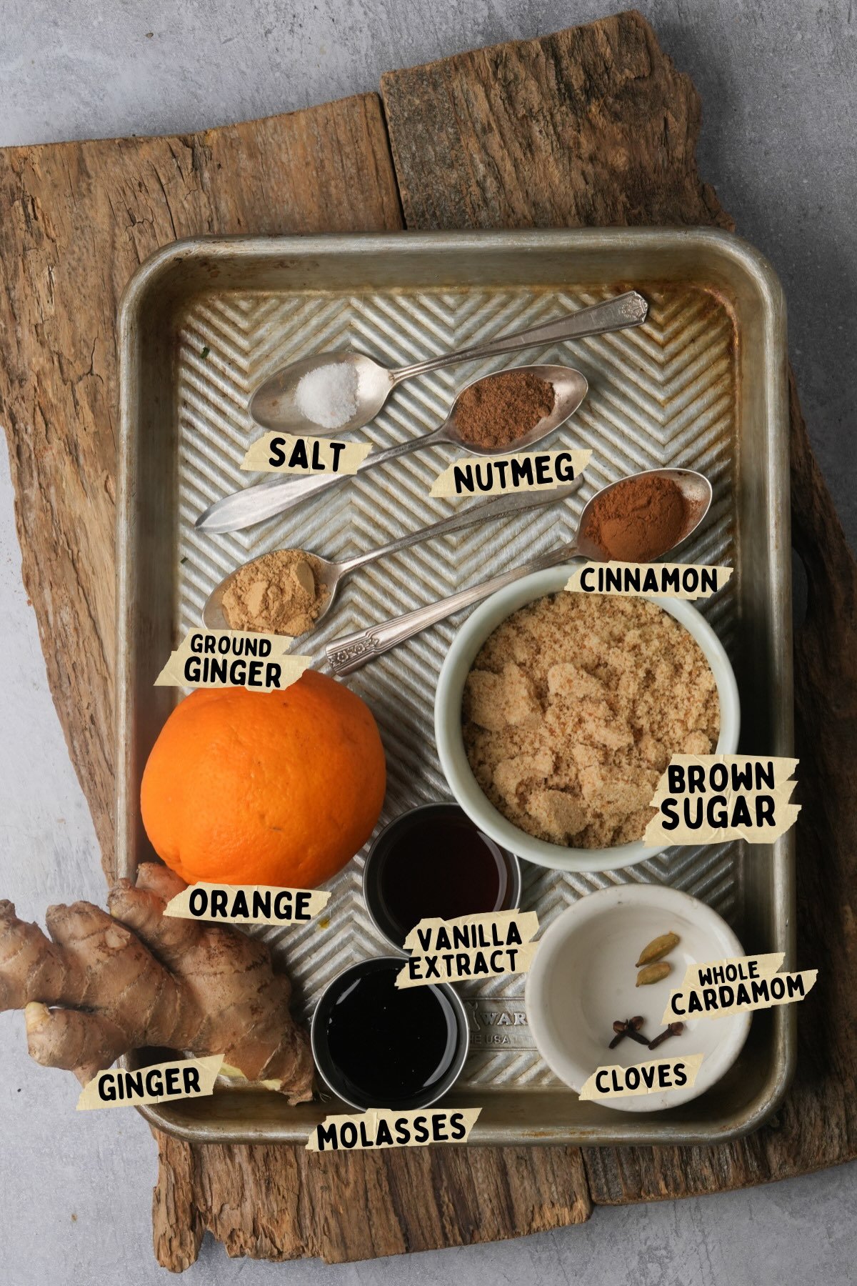 A metal tray on a wooden board holds labeled ingredients for a gingerbread simple syrup recipe: salt, nutmeg, cinnamon, ground ginger, an orange, brown sugar, ginger root, vanilla extract, molasses, whole cardamom, and cloves.