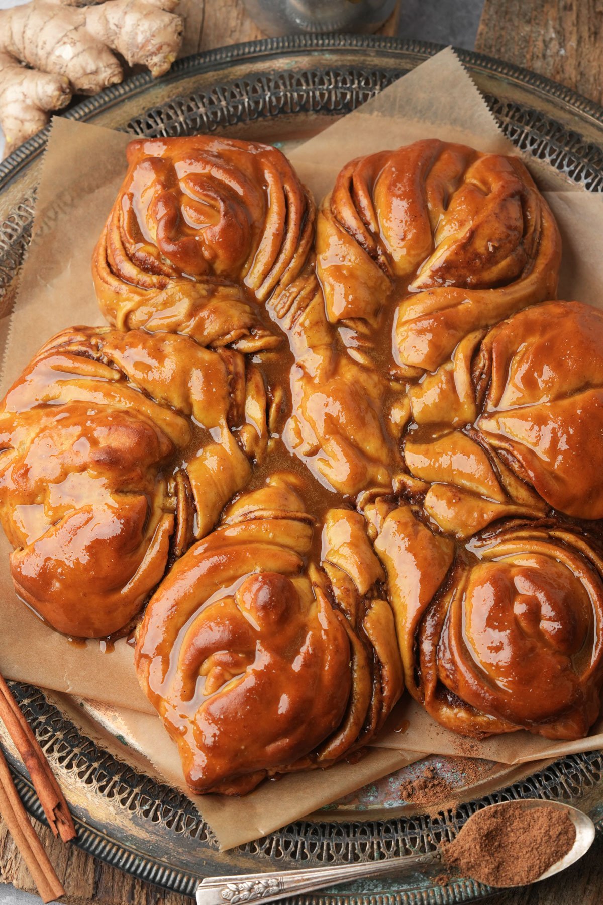 A close-up of a shiny, golden-brown pull-apart gingerbread star bread on parchment paper, arranged in a flower pattern on a decorative silver tray, with cinnamon sticks and fresh ginger nearby.