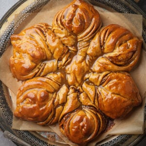 A glossy, gingerbread star bread with swirled layers sits on parchment paper atop a decorative metal tray. The pastry appears freshly baked and coated with a shiny glaze.