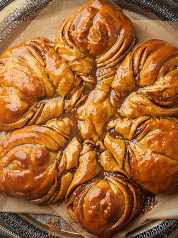 A glossy, gingerbread star bread with swirled layers sits on parchment paper atop a decorative metal tray. The pastry appears freshly baked and coated with a shiny glaze.