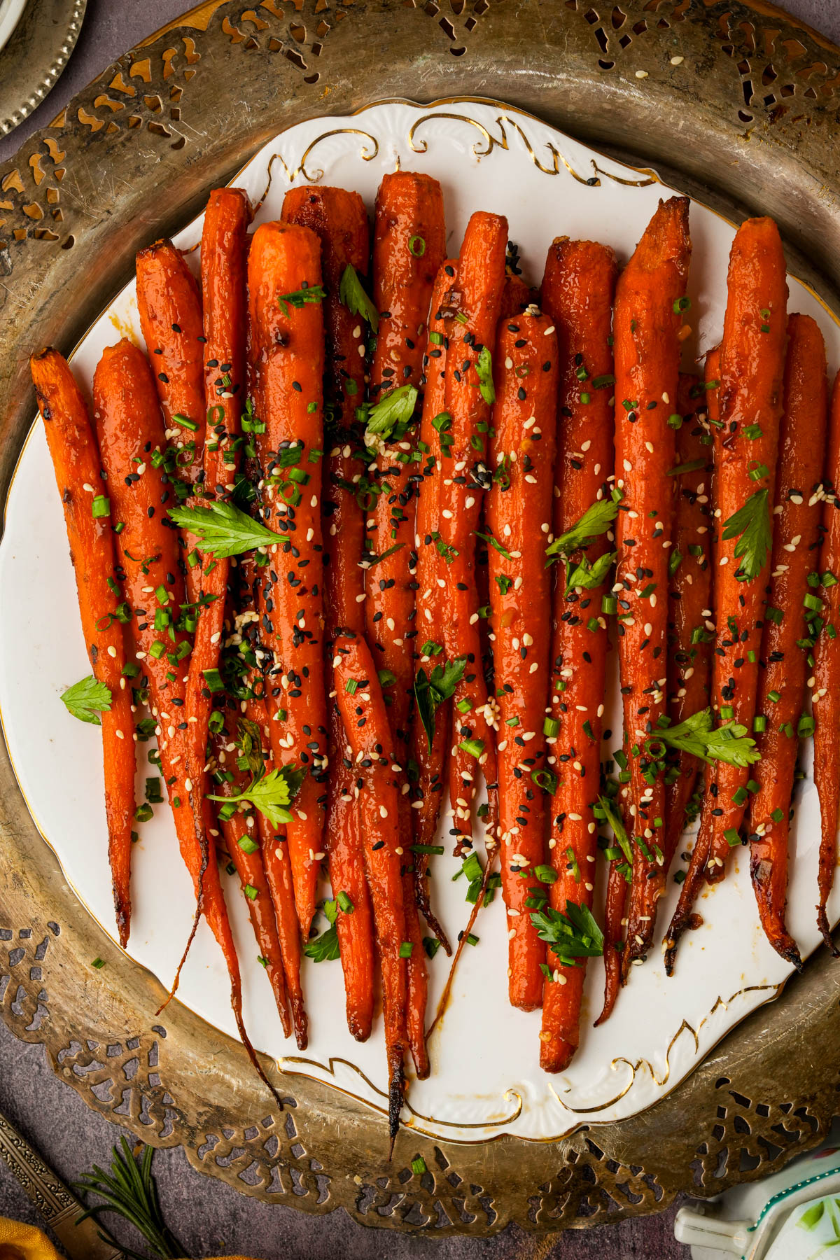 Whole maple roasted carrots arranged on a decorative white plate, garnished with chopped herbs, sesame seeds, and seasoning, set on an ornate metal tray.