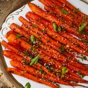 A plate of maple roasted carrots garnished with sesame seeds, chopped chives, and fresh parsley, arranged neatly on a decorative white platter.
