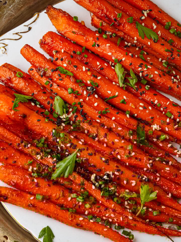 A plate of maple roasted carrots garnished with sesame seeds, chopped chives, and fresh parsley, arranged neatly on a decorative white platter.