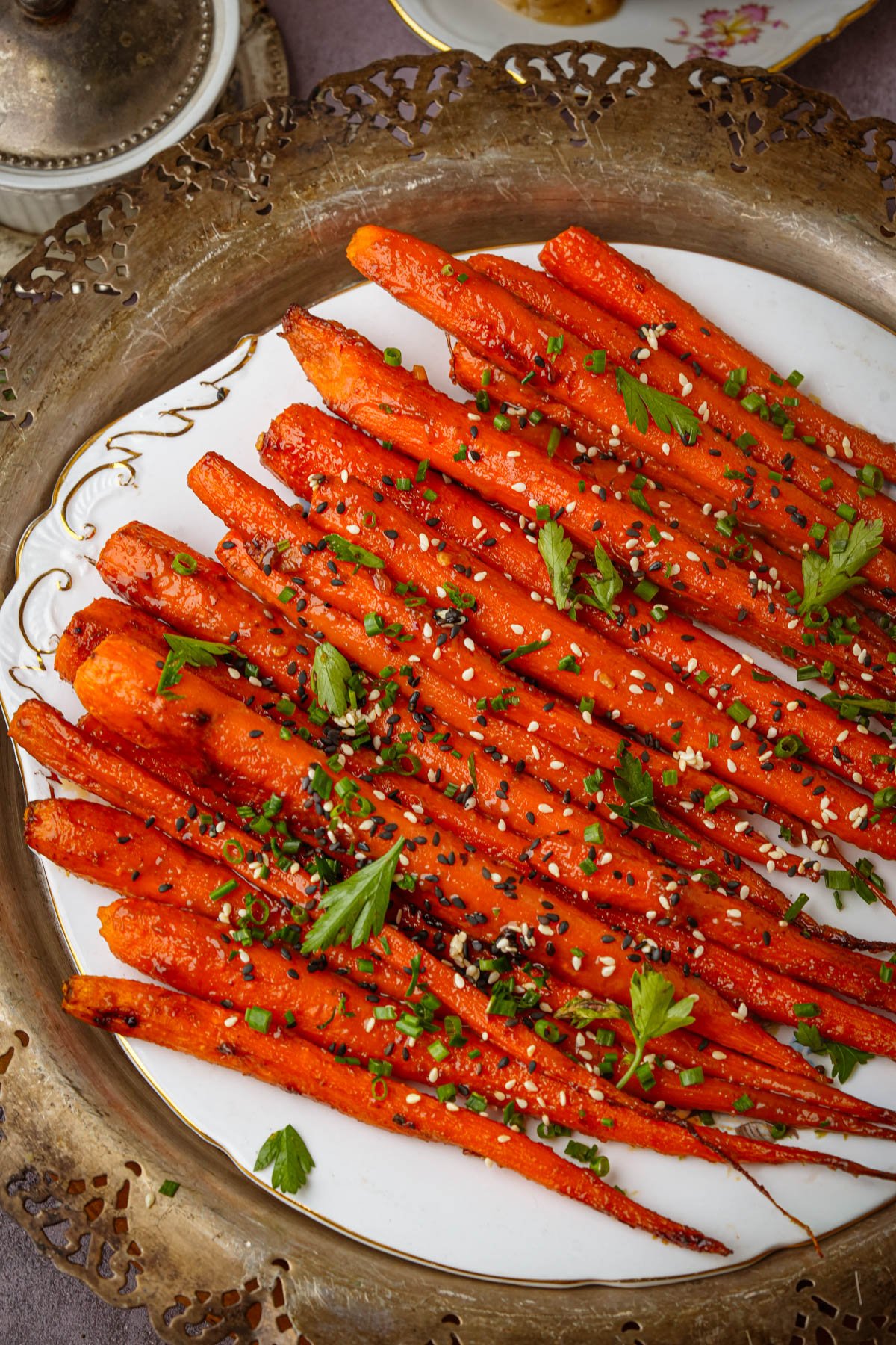 Whole maple roasted carrots arranged on a white plate, garnished with chopped chives, parsley, and sprinkled with black and white sesame seeds, served on an ornate metallic tray.