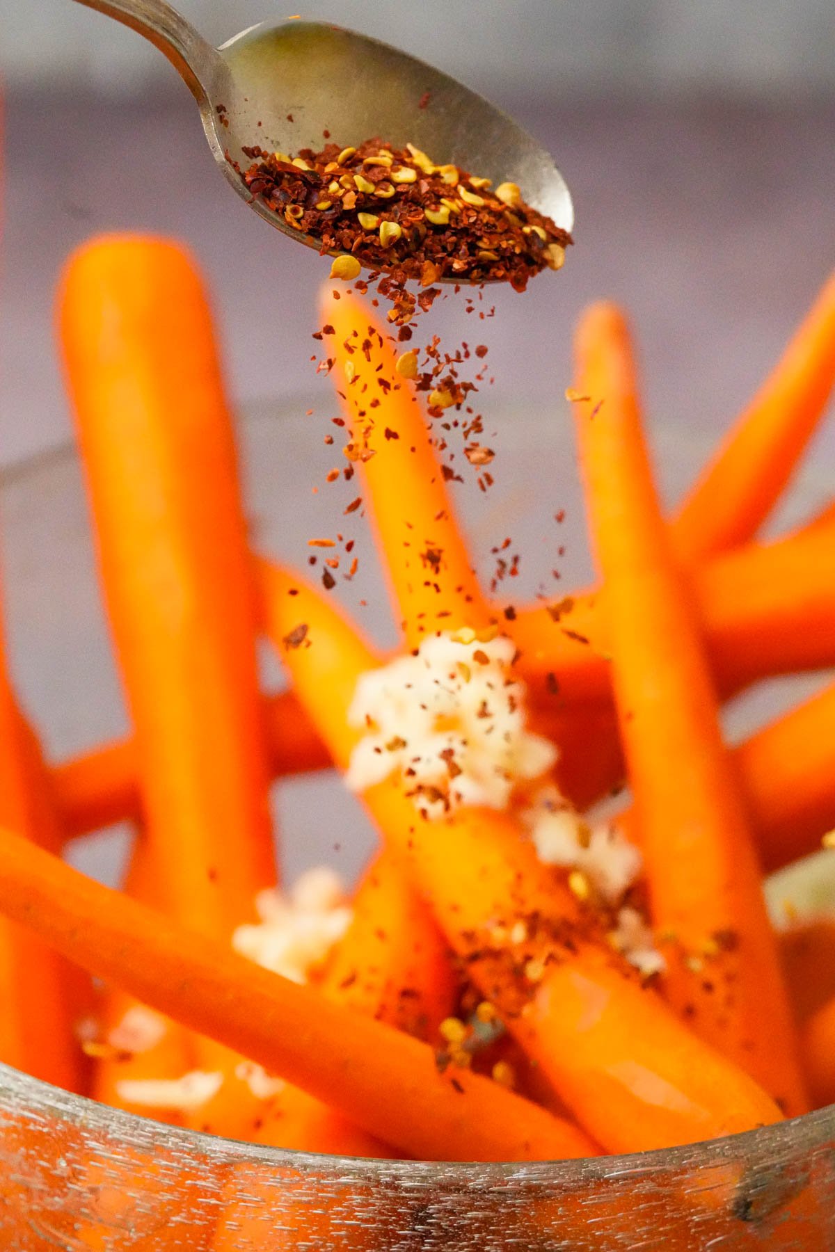 A spoon sprinkles red chili flakes over a bowl of whole peeled carrots and chopped garlic, preparing the vegetables for seasoning.