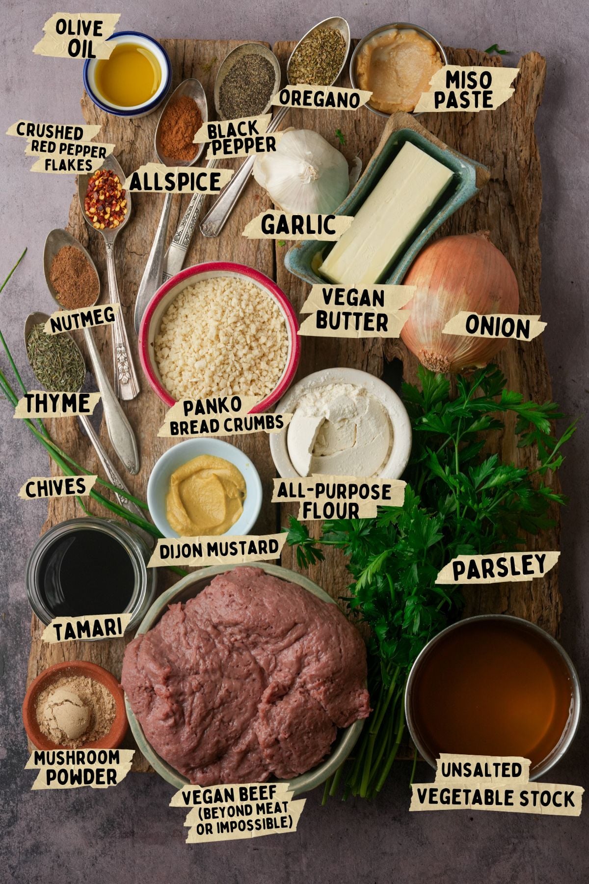 A wooden board topped with labeled ingredients for vegan Swedish meatballs, including vegan ground beef, panko, onion, garlic, vegan butter, flour, spices, herbs, vegetable stock, tamari, mustard, miso paste, and olive oil.