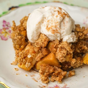 A serving of vegan apple crisp topped with a scoop of vanilla ice cream and sprinkled with cinnamon, presented on a decorative floral plate.