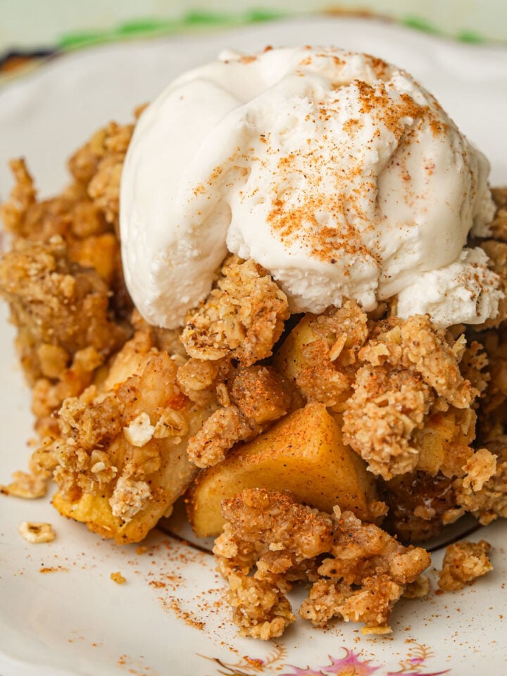 A serving of vegan apple crisp topped with a scoop of vanilla ice cream and sprinkled with cinnamon, presented on a decorative floral plate.