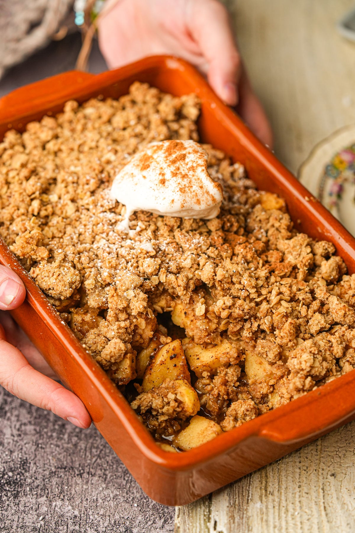 A person holding a rectangular orange baking dish filled with vegan apple crumble, topped with a dollop of whipped cream and a sprinkle of cinnamon. The dessert sits on a rustic wooden table.