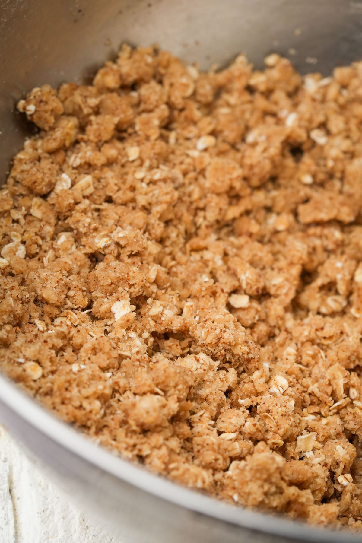 Close-up of a metal mixing bowl filled with a crumbly oat and brown sugar mixture, likely used as a topping for baked goods such as crisps or crumbles.