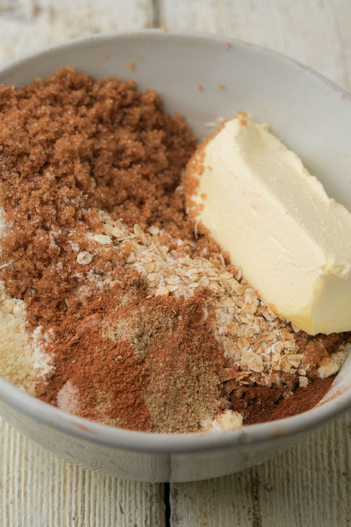 A bowl contains brown sugar, a block of butter, oats, flour, and various spices, all measured and unmixed, on a light wooden surface.