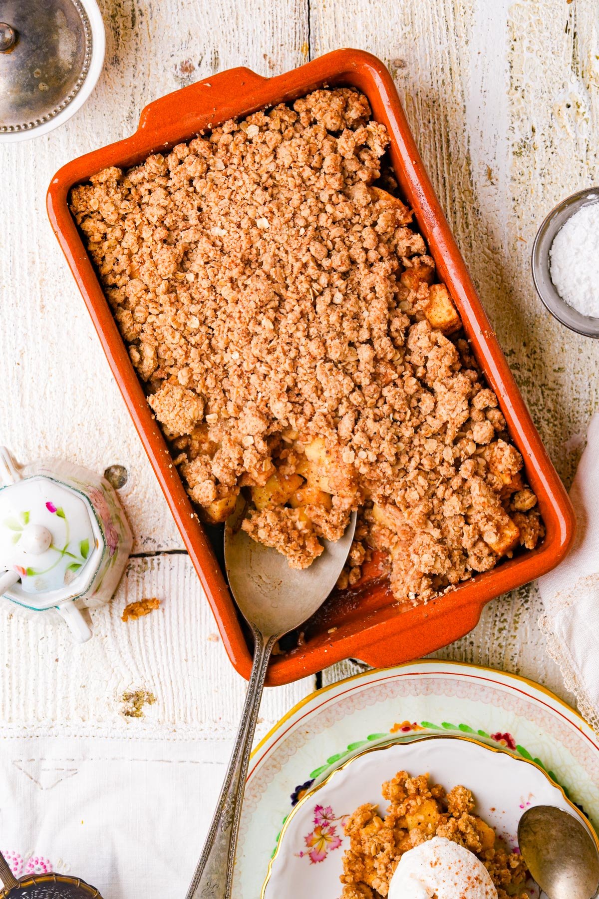 A rectangular baking dish filled with golden-brown vegan apple crumble, with a serving spoon scooping some out. Nearby are a plate with a portion of crumble and a scoop of ice cream, a sugar bowl, and a small metal dish.
