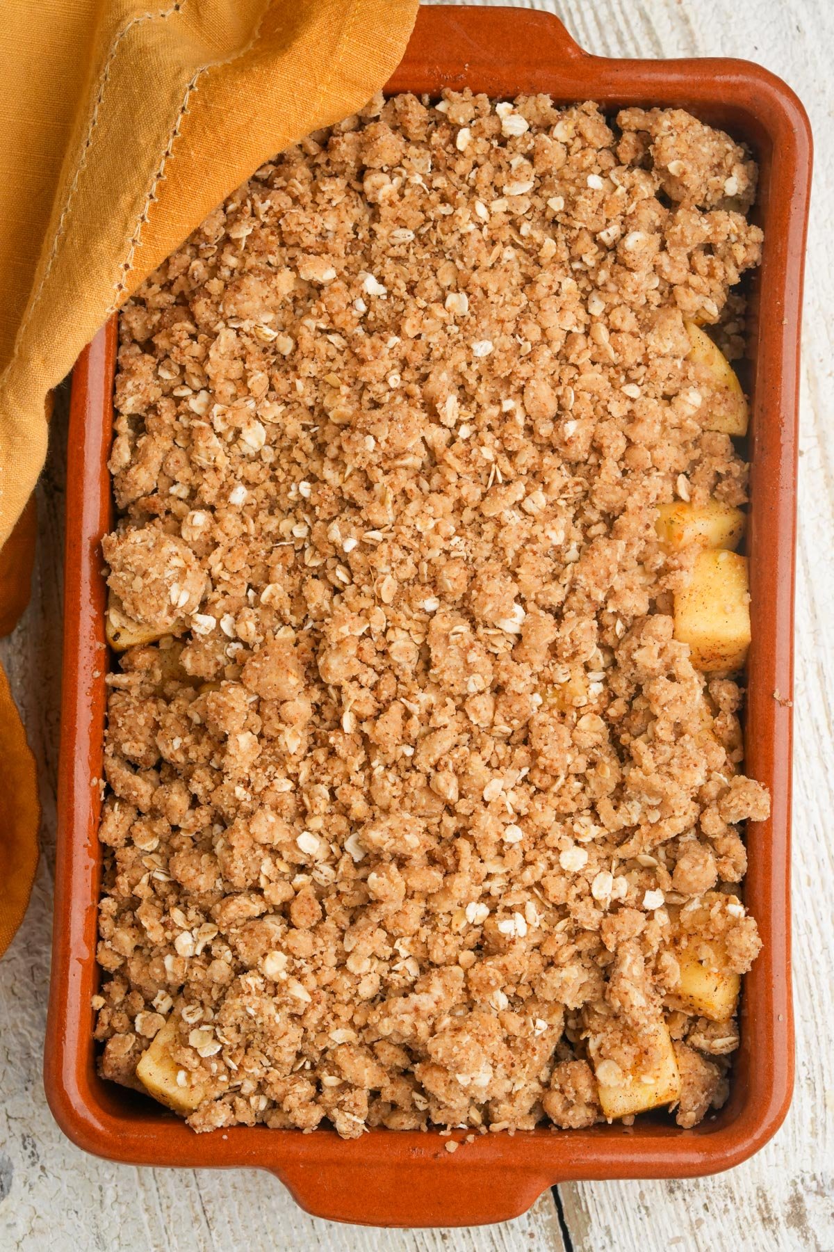 A rectangular baking dish filled with apple crisp, topped with a golden-brown oat and cinnamon crumble. A tan cloth is partially draped over one corner of the dish, which sits on a light wooden surface.