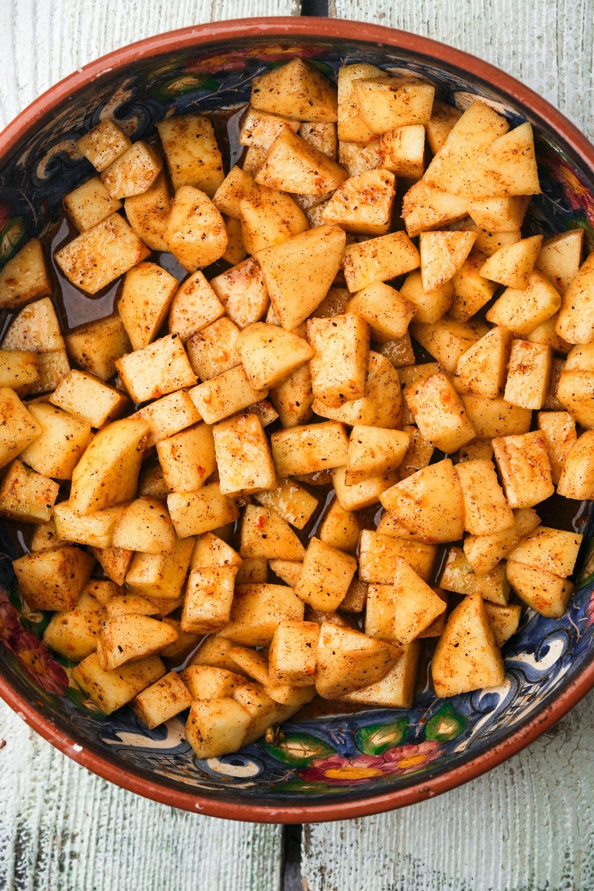 A bowl filled with chopped apples coated in cinnamon and sugar, placed on a rustic white wooden surface. The apples are evenly diced and thoroughly mixed.