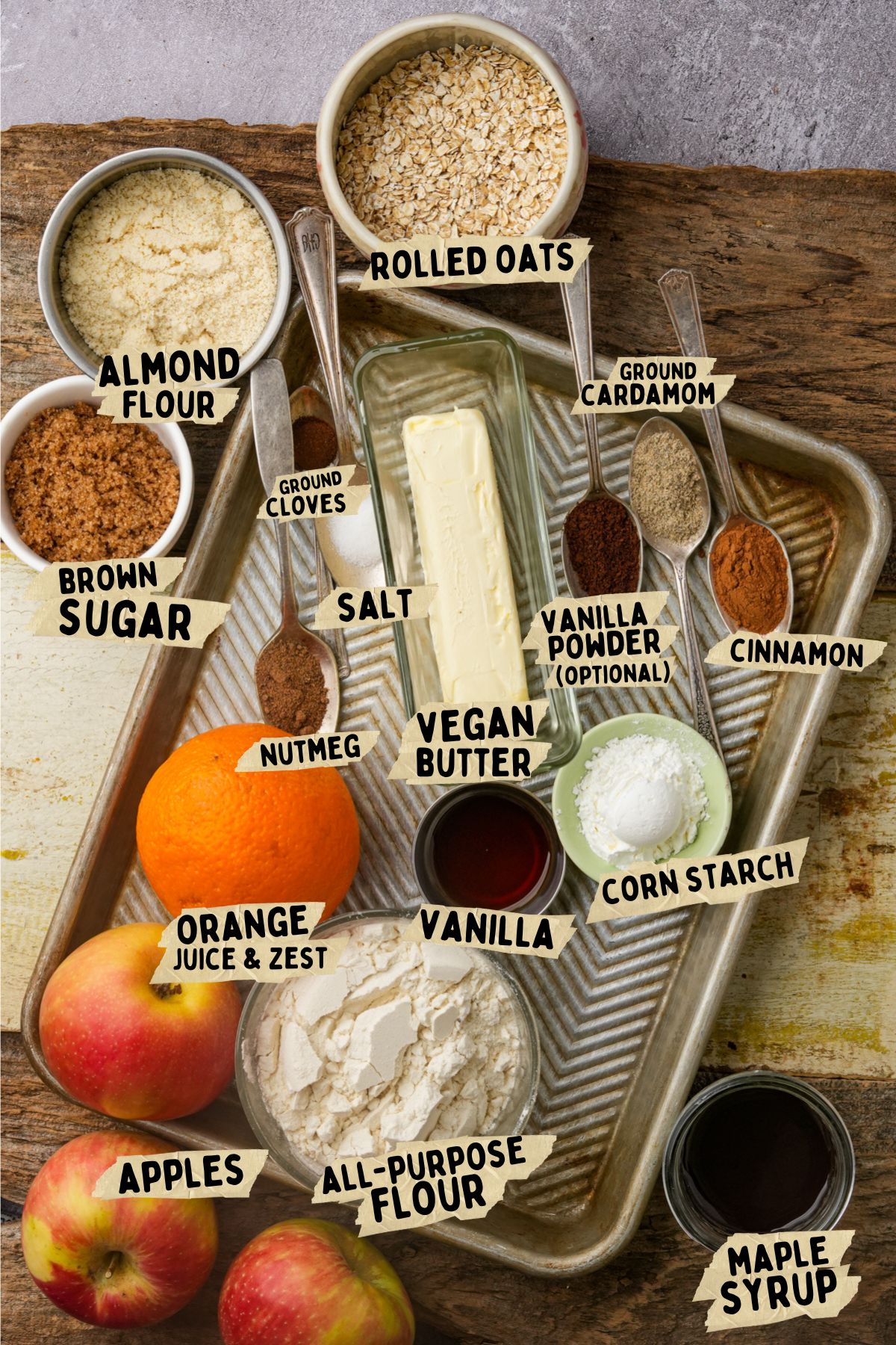 A baking tray holds labeled ingredients for baking: rolled oats, ground cardamom, almond flour, ground cloves, salt, vegan butter, brown sugar, vanilla powder, cinnamon, nutmeg, orange juice and zest, vanilla, corn starch, apples, all-purpose flour, and maple syrup.