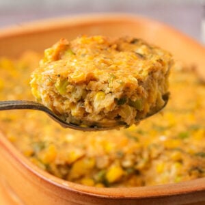 A spoonful of vegan corn casserole held above a baking dish filled with more of the casserole, showing a moist, crumbly texture with visible corn and green herbs.
