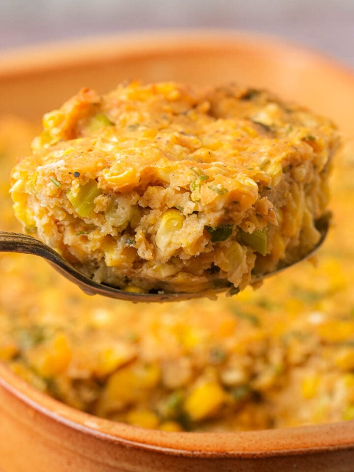 A spoonful of vegan corn casserole held above a baking dish filled with more of the casserole, showing a moist, crumbly texture with visible corn and green herbs.