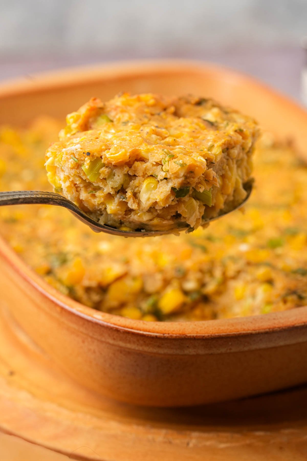 A spoon lifting a serving of vegan corn casserole from a brown baking dish, showing a moist, golden, and textured mixture with visible pieces of corn and herbs.