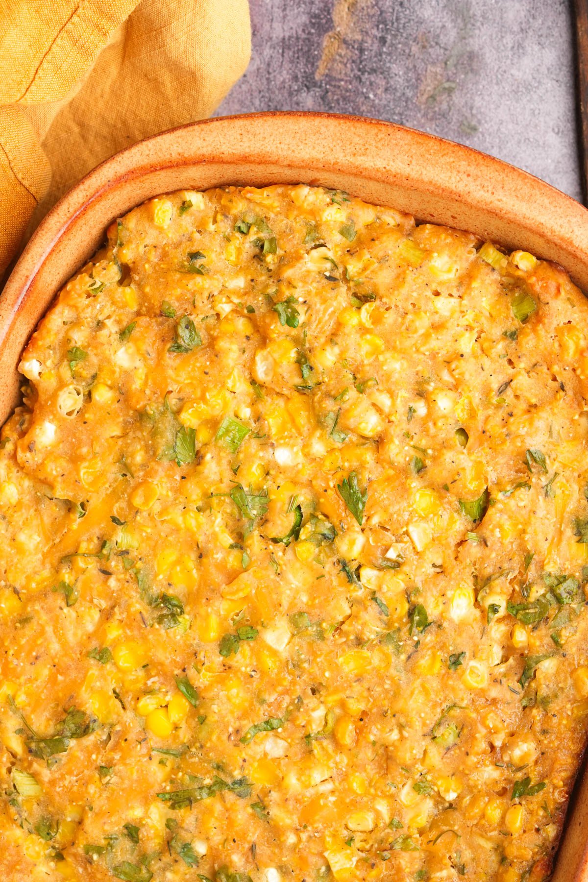 A baked cheesy vegan corn casserole in a brown dish, with visible corn kernels, chopped green herbs, and a golden crust, set on a rustic surface next to a yellow cloth.