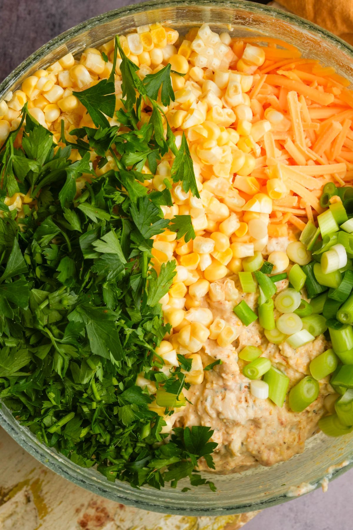 A glass bowl filled with chopped fresh parsley, shredded cheddar cheese, corn kernels, sliced green onions, and vegan corn casserole batter, all arranged in separate sections.