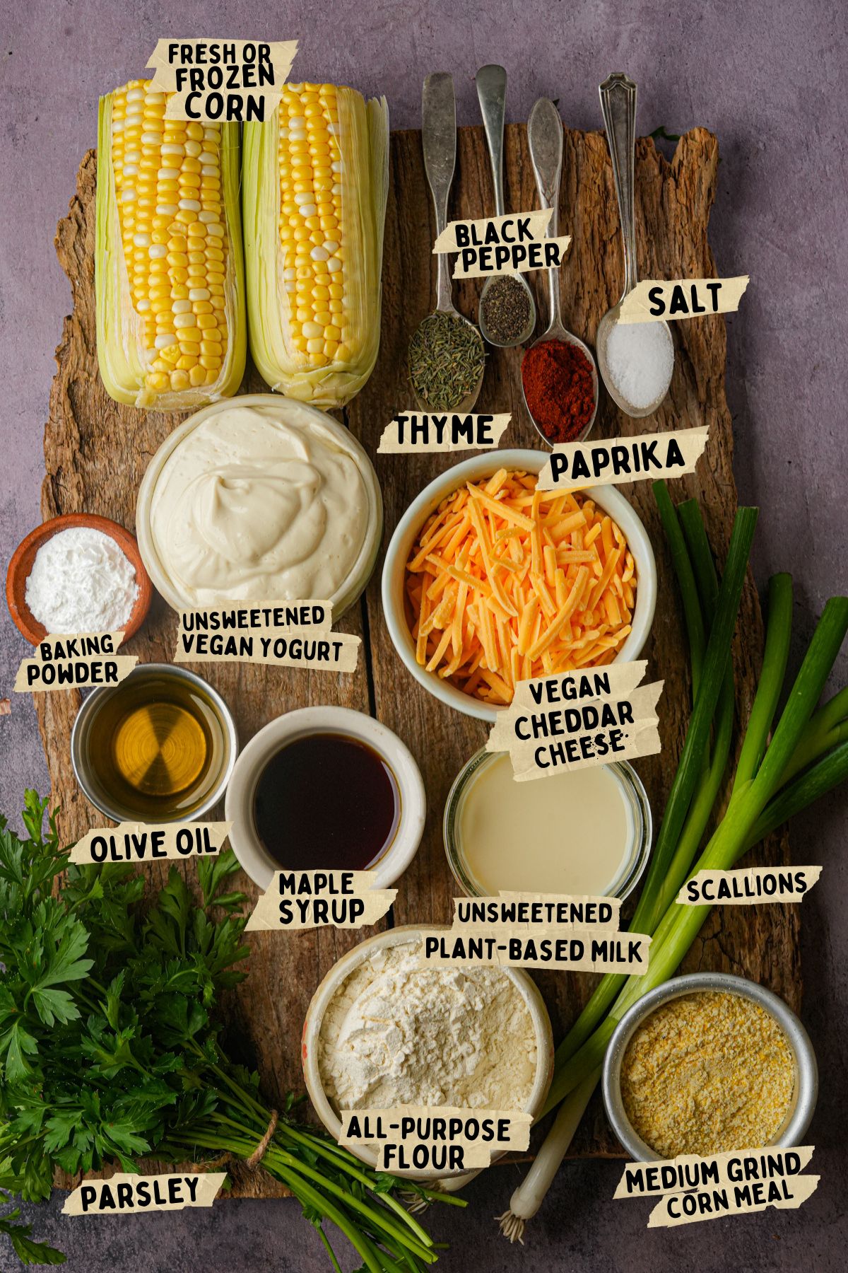 A flat lay of ingredients on a wooden board for a vegan corn casserole recipe, including corn, vegan yogurt, cheddar cheese, plant-based milk, cornmeal, flour, olive oil, maple syrup, scallions, parsley, baking powder, salt, pepper, thyme, and paprika.