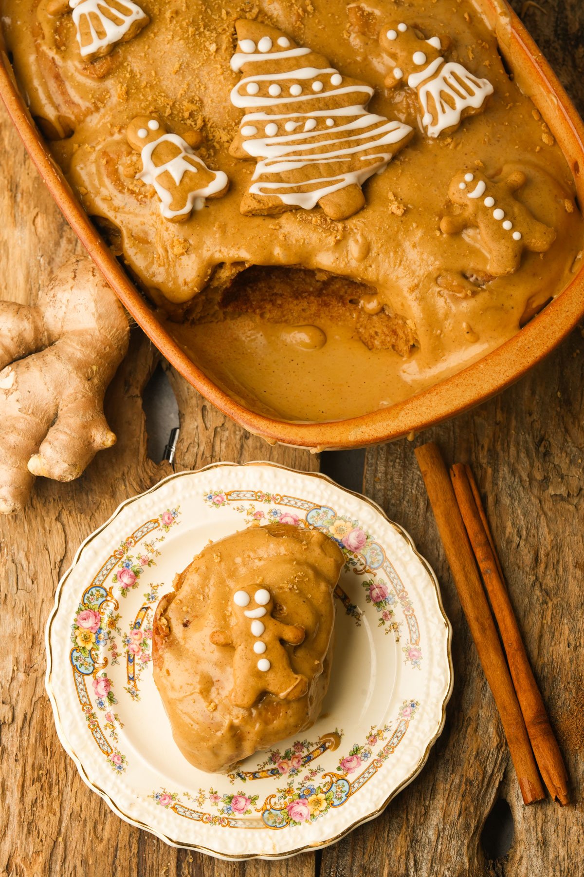 A vegan gingerbread cinnamon roll with brown icing is served on a decorative plate, while the rest of the rolls are in a baking dish, topped with icing-decorated cookie shapes. Ginger and cinnamon sticks are nearby on a rustic wooden surface.