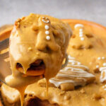 A close-up of Gingerbread Cinnamon Rolls covered in caramel icing and topped with a small gingerbread man cookie, with more decorated rolls blurred in the background.