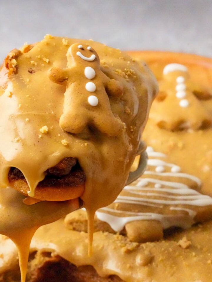 A close-up of Gingerbread Cinnamon Rolls covered in caramel icing and topped with a small gingerbread man cookie, with more decorated rolls blurred in the background.