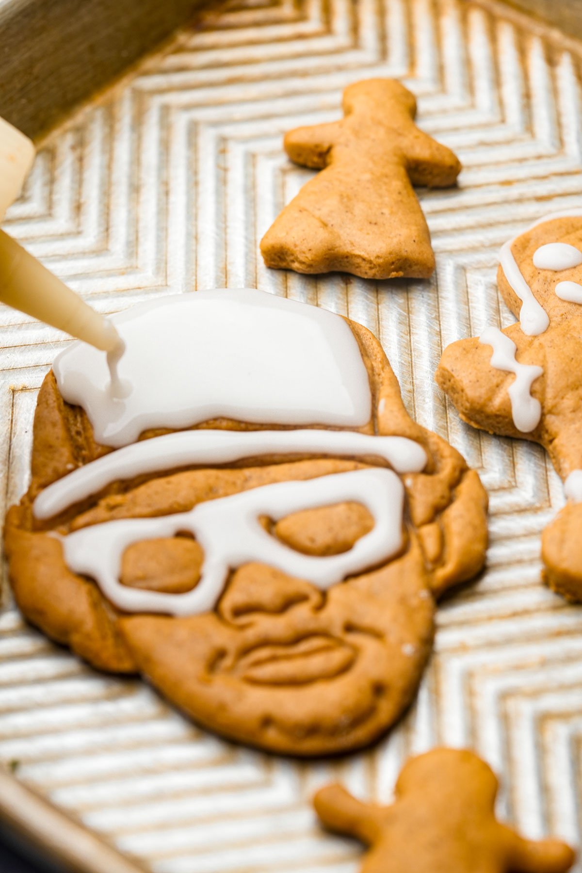 Vegan gingerbread cookies shaped like Eazy-E wearing glasses and a hat, being decorated with white icing on a baking tray. Other small gingerbread cookies are nearby.