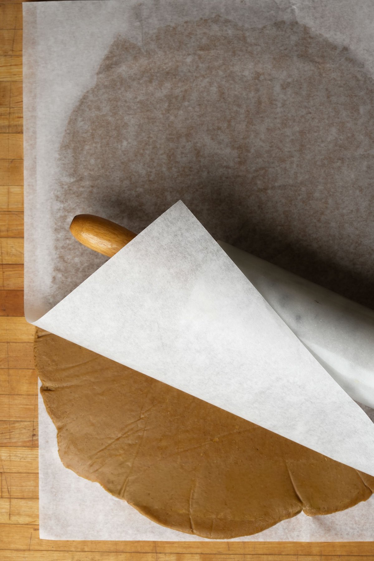 A sheet of vegan gingerbread cookie dough is being rolled out between two pieces of parchment paper with a rolling pin on a wooden surface. The top layer of parchment is partially lifted to reveal the dough underneath.