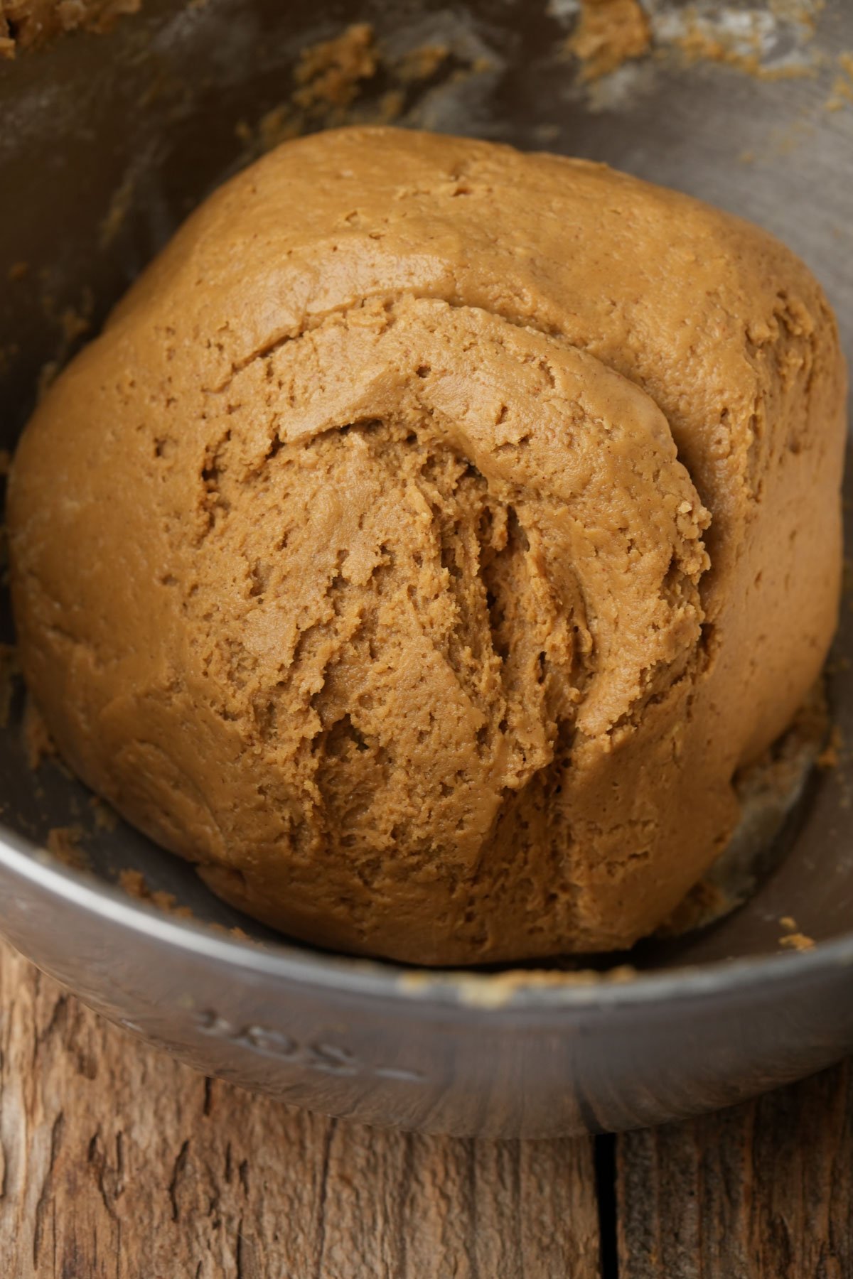 A close-up of a ball of brown vegan gingerbread cookie dough in a metal mixing bowl, resting on a rustic wooden surface. The dough has a slightly rough, textured surface and appears freshly mixed.