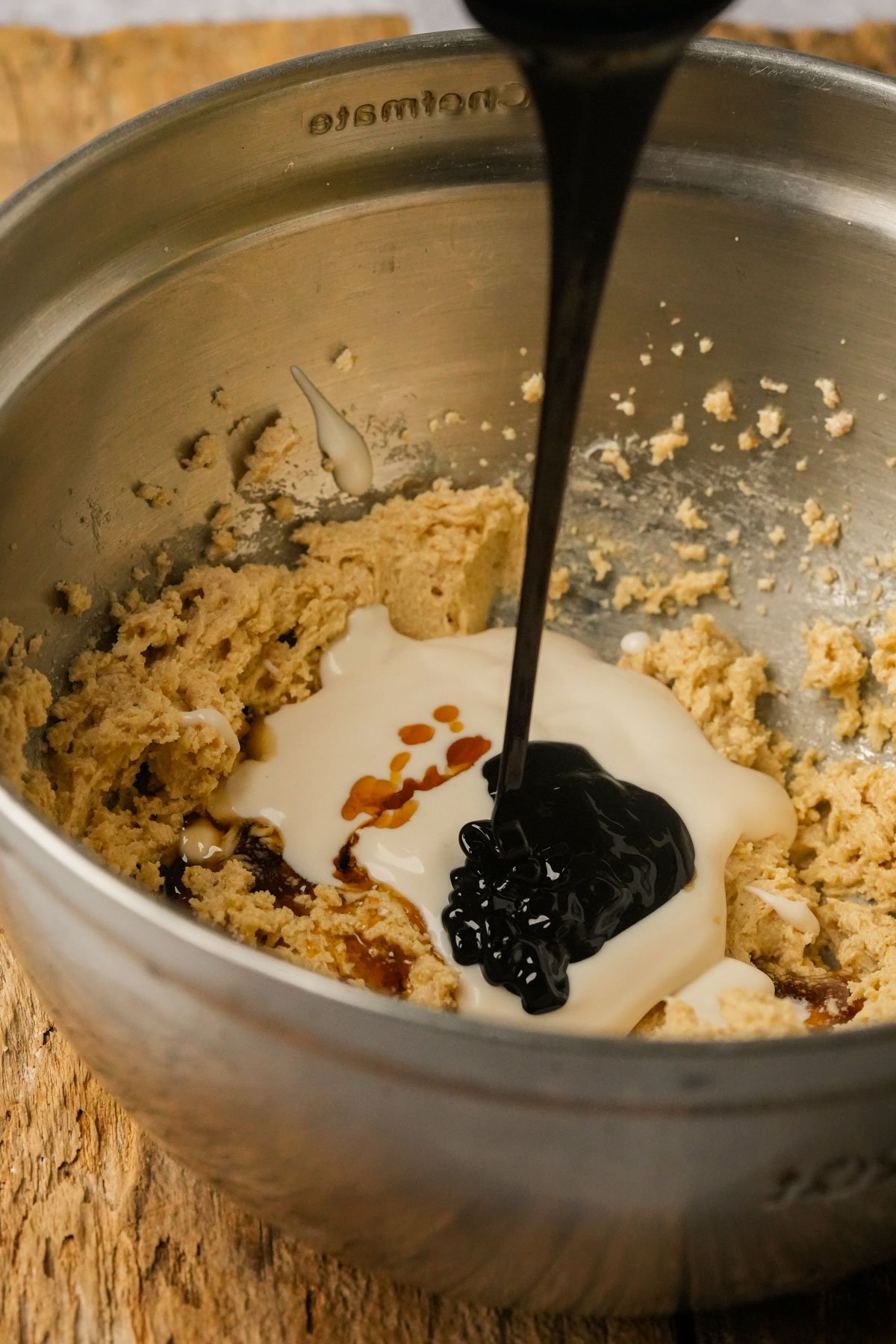 A close-up of a metal mixing bowl with partially mixed vegan gingerbread cookie dough. Thick black syrup and creamy liquid are being poured onto the mixture, which sits on a wooden surface.