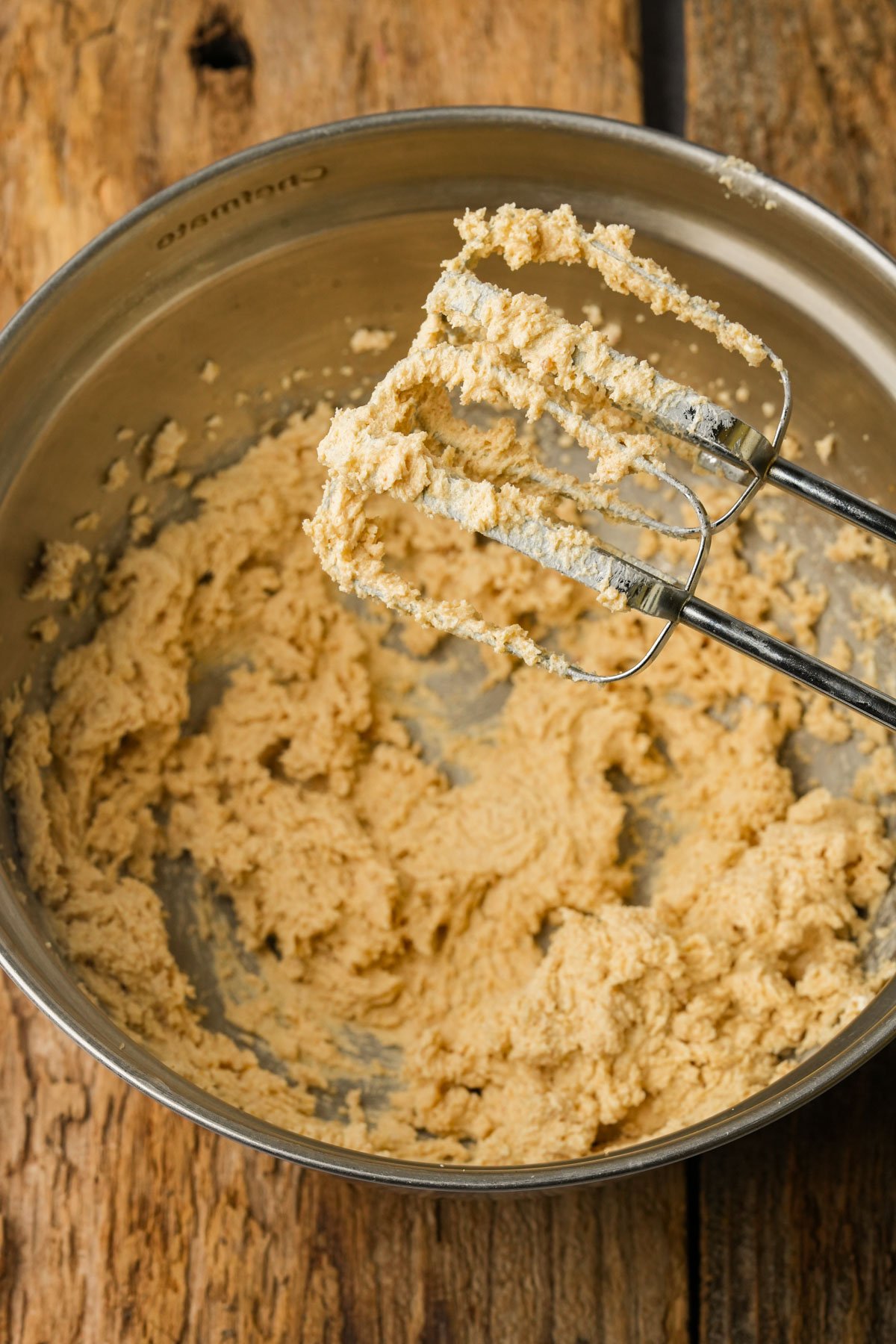 A metal mixing bowl with creamy, light brown whipped vegan butter mixture inside sits on a wooden surface. An electric mixer with batter on its beaters rests above the bowl.