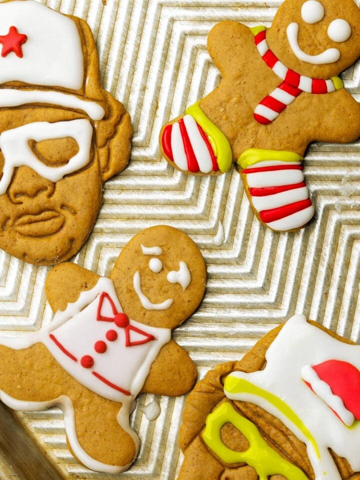 Vegan gingerbread cookies decorated with colorful icing on a baking sheet; two gingerbread people with red, white, and yellow details, and two cookies shaped like faces wearing hats and glasses.