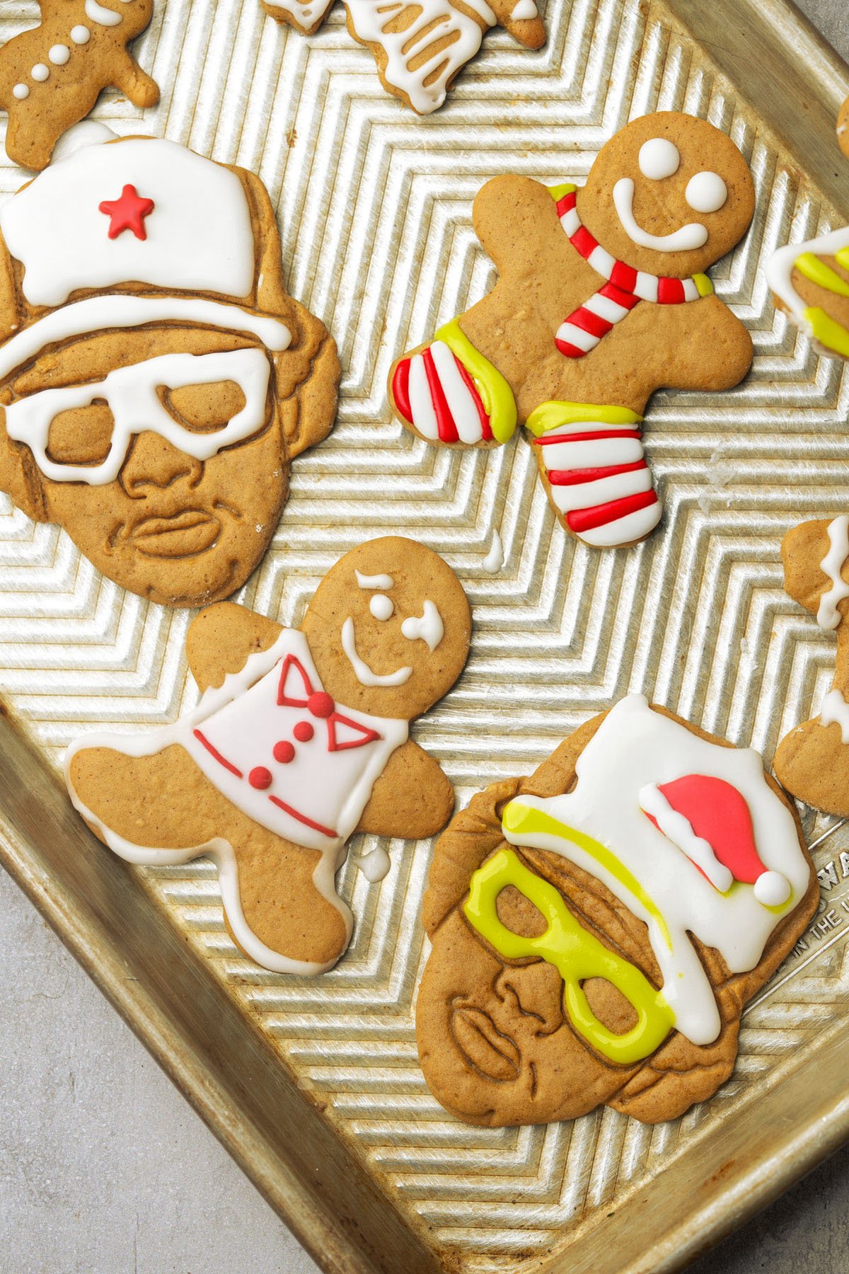 A tray of decorated vegan gingerbread cookies, including classic gingerbread people and cookies shaped like faces wearing glasses and hats, all iced with colorful holiday designs.