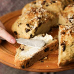 A hand spreads butter on a thick slice of oat and raisin bread using a butter knife. The bread is on a wooden plate with more slices visible in the background.