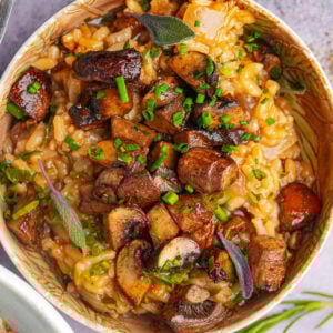 A bowl of creamy mushroom risotto topped with sautéed mushrooms, fresh chives, and sage leaves, garnished with herbs. The dish appears rich and flavorful with a mix of golden and brown hues.