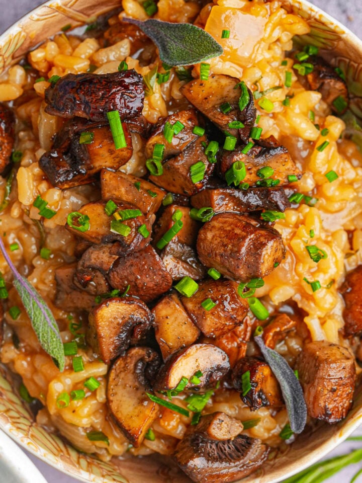 A bowl of creamy mushroom risotto topped with sautéed mushrooms, fresh chives, and sage leaves, garnished with herbs. The dish appears rich and flavorful with a mix of golden and brown hues.