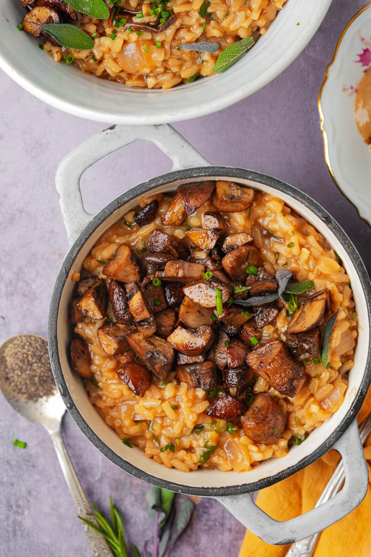 A white pot filled with creamy mushroom risotto topped with sautéed mushrooms and herbs sits on a purple surface, with a spoon, fresh herbs, and a partially visible bowl of risotto nearby.