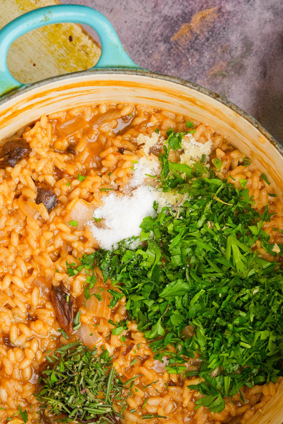 A pot with cooked mushroom risotto, topped with fresh chopped parsley, rosemary, and salt, ready to be mixed. The dish has a rich orange color and is set on a rustic surface.