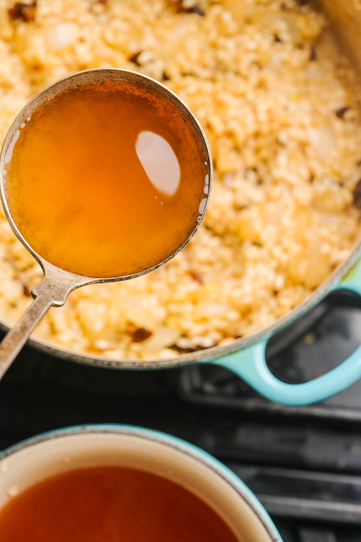 A metal ladle filled with clear brown broth is held over a pot of mushroom risotto, with another pot of broth visible below on a stovetop.