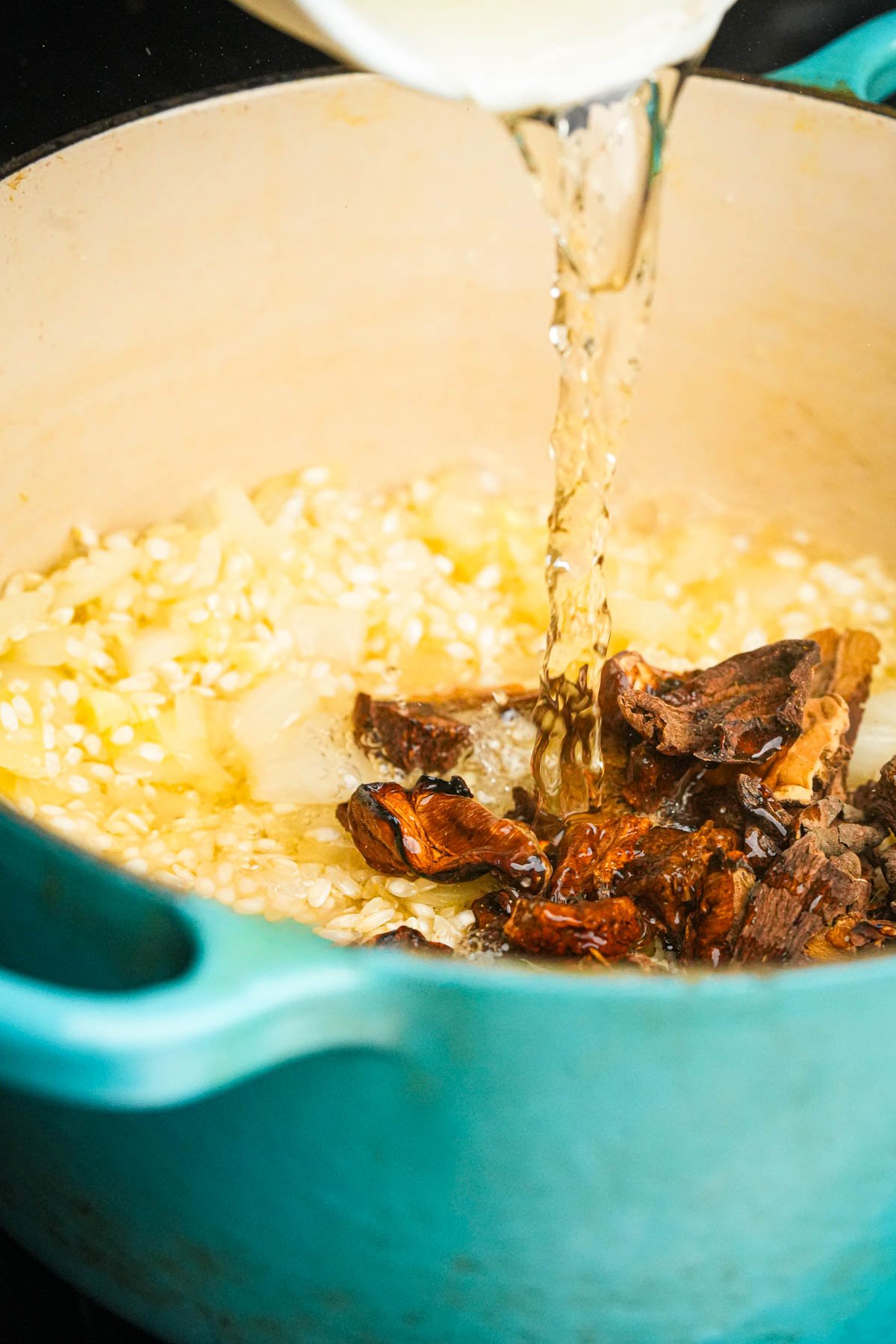 A close-up of a teal pot with onions, rice, dried mushrooms, and wine being poured in, likely the start of a mushroom risotto.