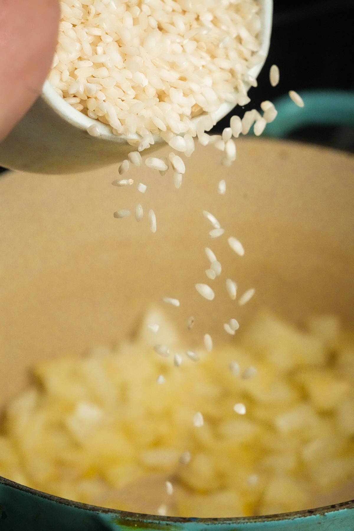 Close-up of uncooked Arborio rice being poured from a white cup into a pot containing chopped onions.