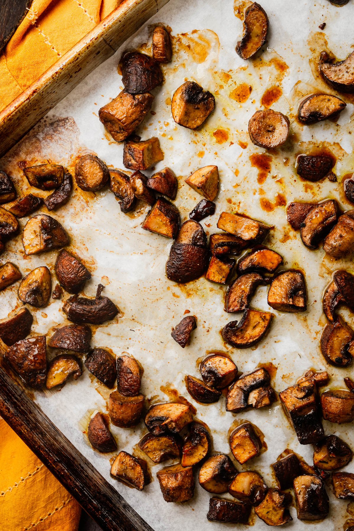 Roasted, sliced mushrooms scattered on a parchment-lined baking sheet, glistening with oil and browned from cooking. An orange cloth napkin is visible in the corner.